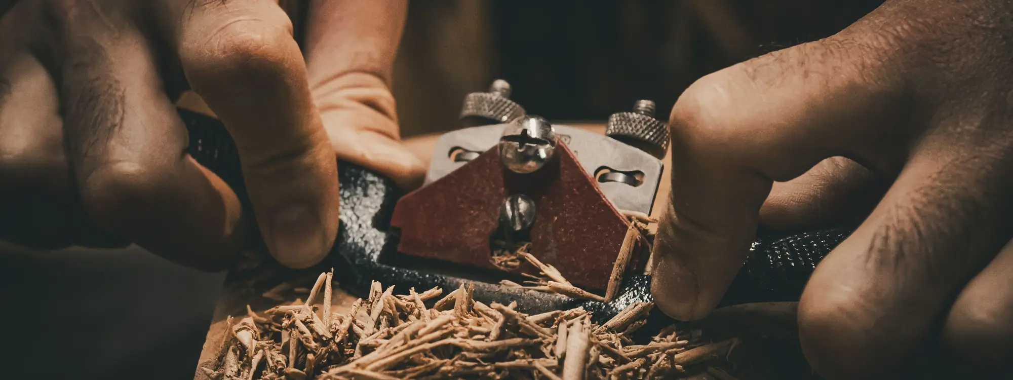 Two hands guiding a hand plane along a board, with a pile of fresh shavings gathering in front of the blade.