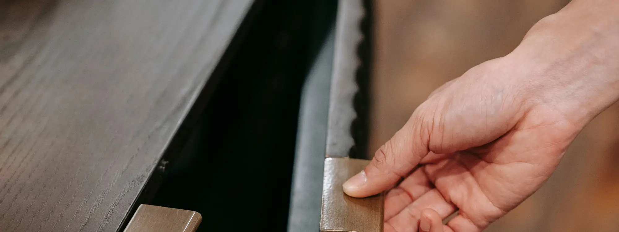 A hand opening a flush cabinet front by a slim aged brass edge pull, with a narrow shadow gap running alongside it.